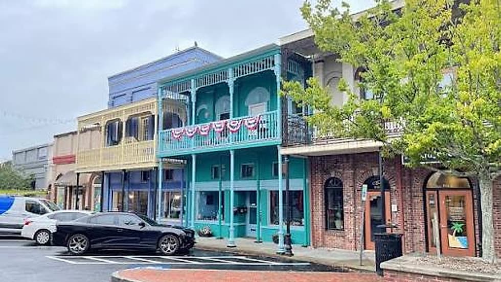 Historic storefronts in downtown Conyers — a teal building with a first-floor balcony and bunting, beside a yellow shopfront, with string lights above the brick street.