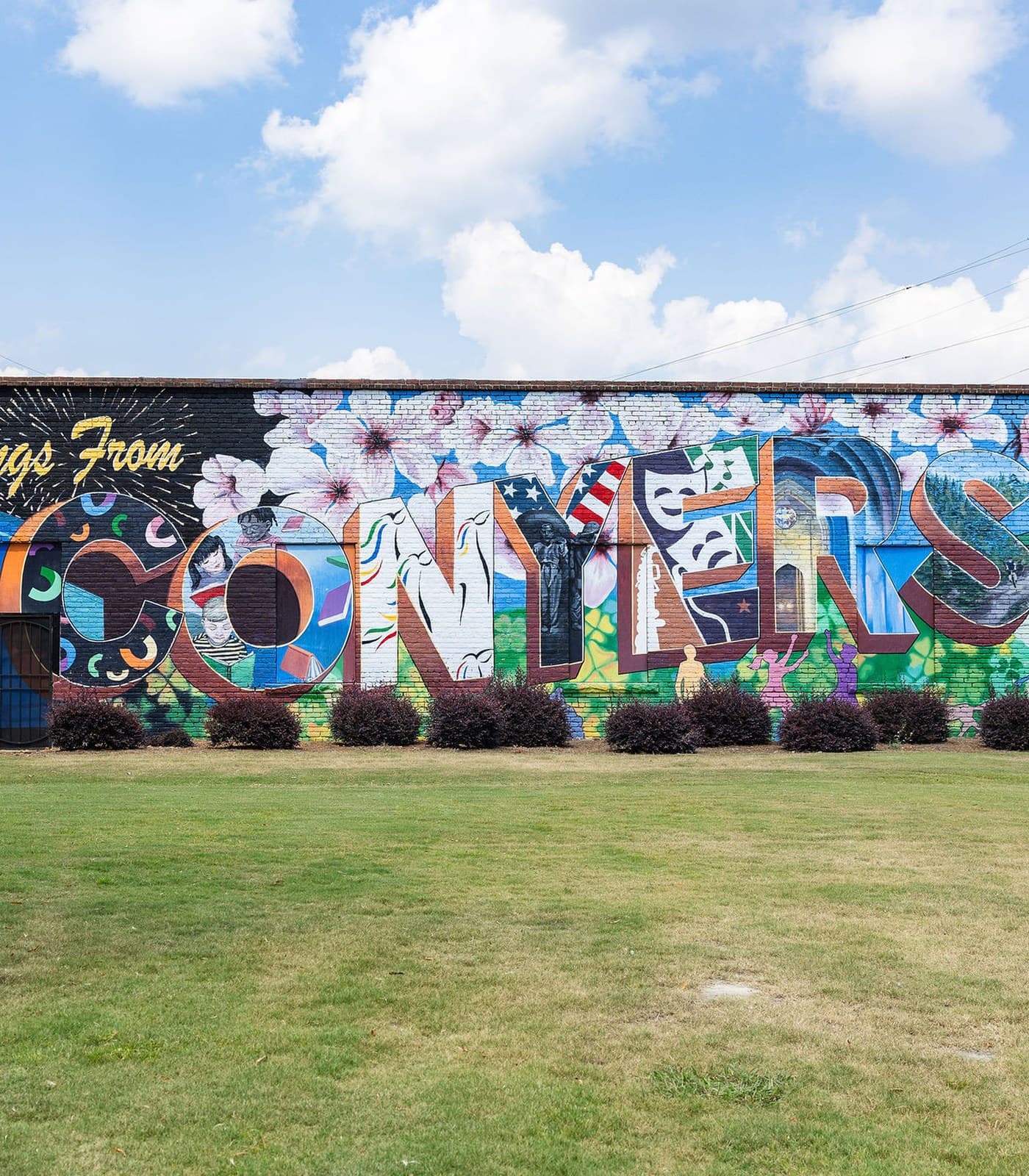 The Greetings From Conyers mural, painted across a brick wall beside a lawn in downtown Conyers.