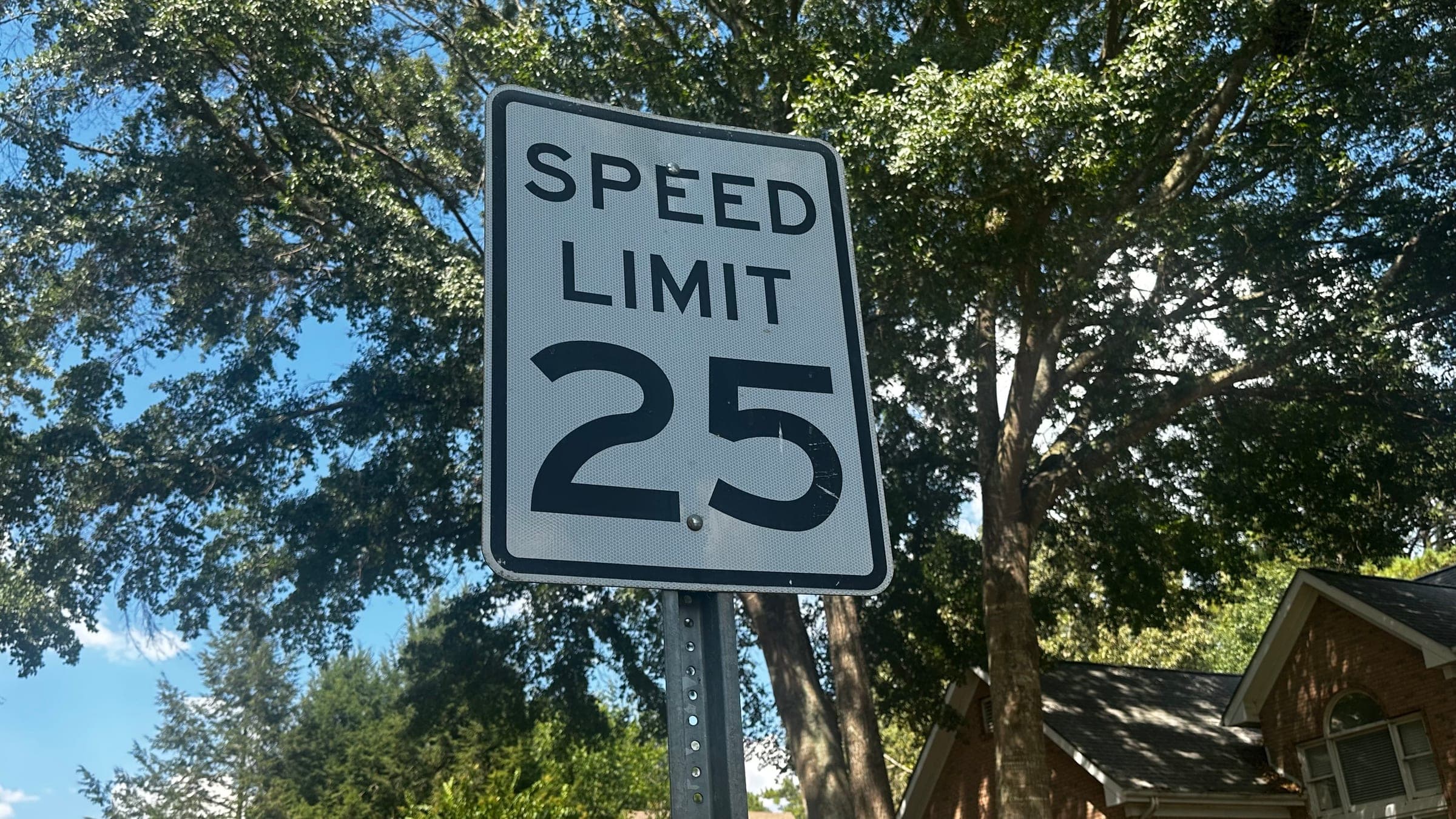 A 25 miles per hour speed limit sign on a tree-lined street in Lambeth Estates, with the roof of a brick home behind it.
